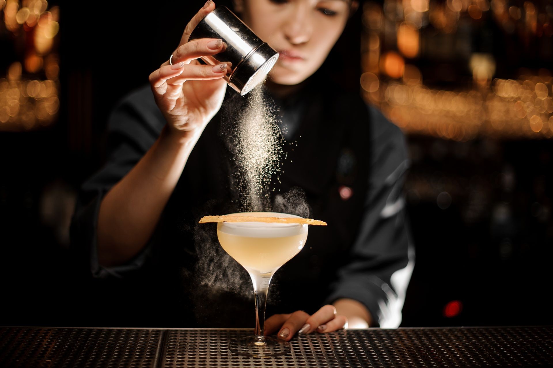 Professional bartender girl holding a spice shaker adding to a delicious cocktail flavours on the bar counter in the blurred background